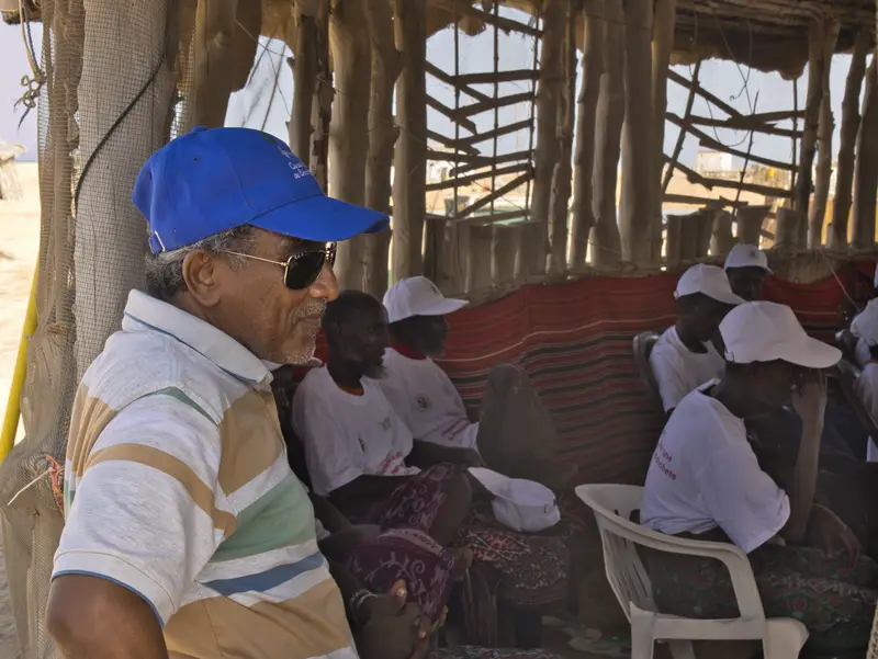 Un ancien en casquette bleue parmi des participants à l’ombre.